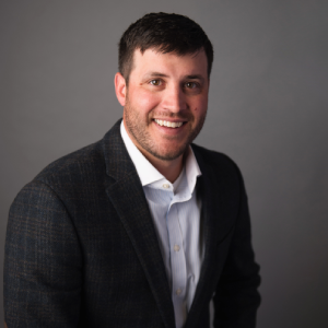 A professional headshot of a smiling man with dark hair and a light beard, wearing a white button-down shirt under a dark, subtly patterned blazer, against a neutral gray background - https://summitstates.com/