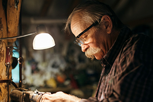 An elderly man with glasses and a white mustache intently focuses on manual work in a dimly lit workshop, illuminated by the warm glow of a desk lamp
