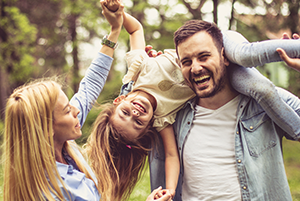 A joyful family plays together outdoors, with a laughing father holding his daughter upside down over his shoulder while the mother smiles and holds her hand
