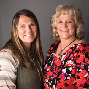 Two women, one younger and one older, are smiling side-by-side in a studio portrait against a solid gray background - https://summitstates.com/